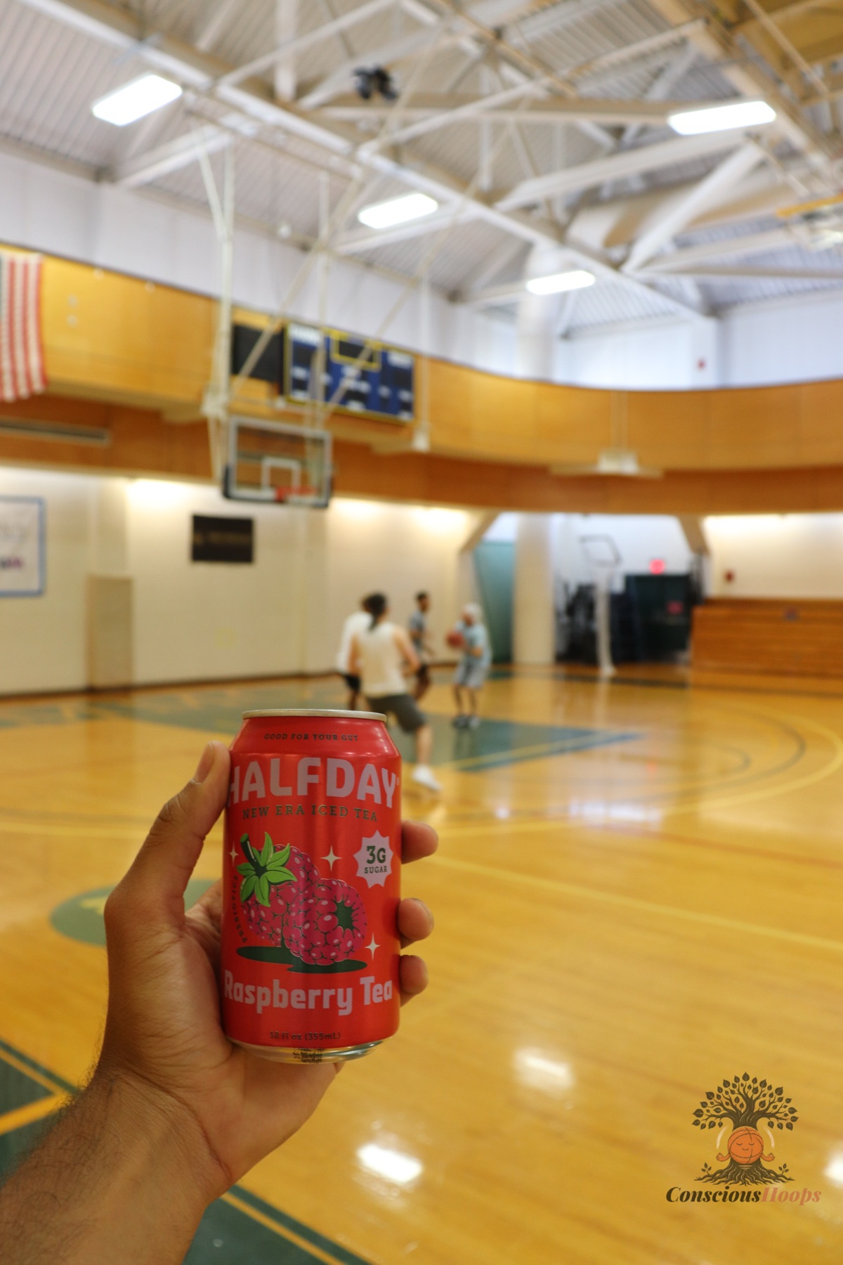 A hand holding a Halfday iced tea can with the gym court in the background